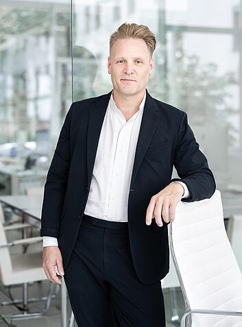 Mann in Anzug steht an einem weißen Stuhl in einem modernen Büro mit Glaswänden.