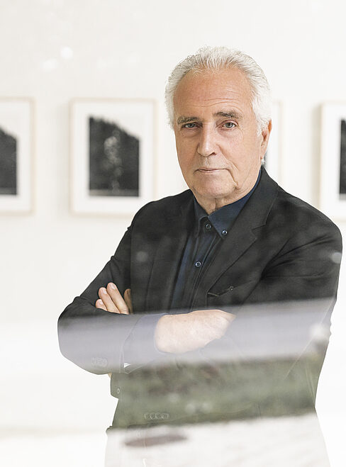 Ein Mann mit weißem Haar steht vor gerahmten Schwarz-Weiß-Fotos in einer Galerie.