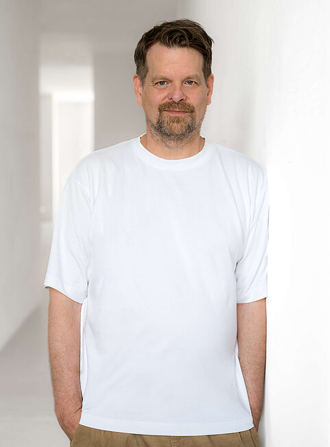 Mann in einem weißen T-Shirt lehnt an einer Wand in einem hellen, minimalistischen Raum.