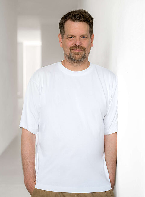 Mann in einem weißen T-Shirt lehnt an einer Wand in einem hellen, minimalistischen Raum.
