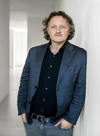 Nachdenklicher Mann mit Locken und dunkel blauem Sakko lehnt an heller Wand