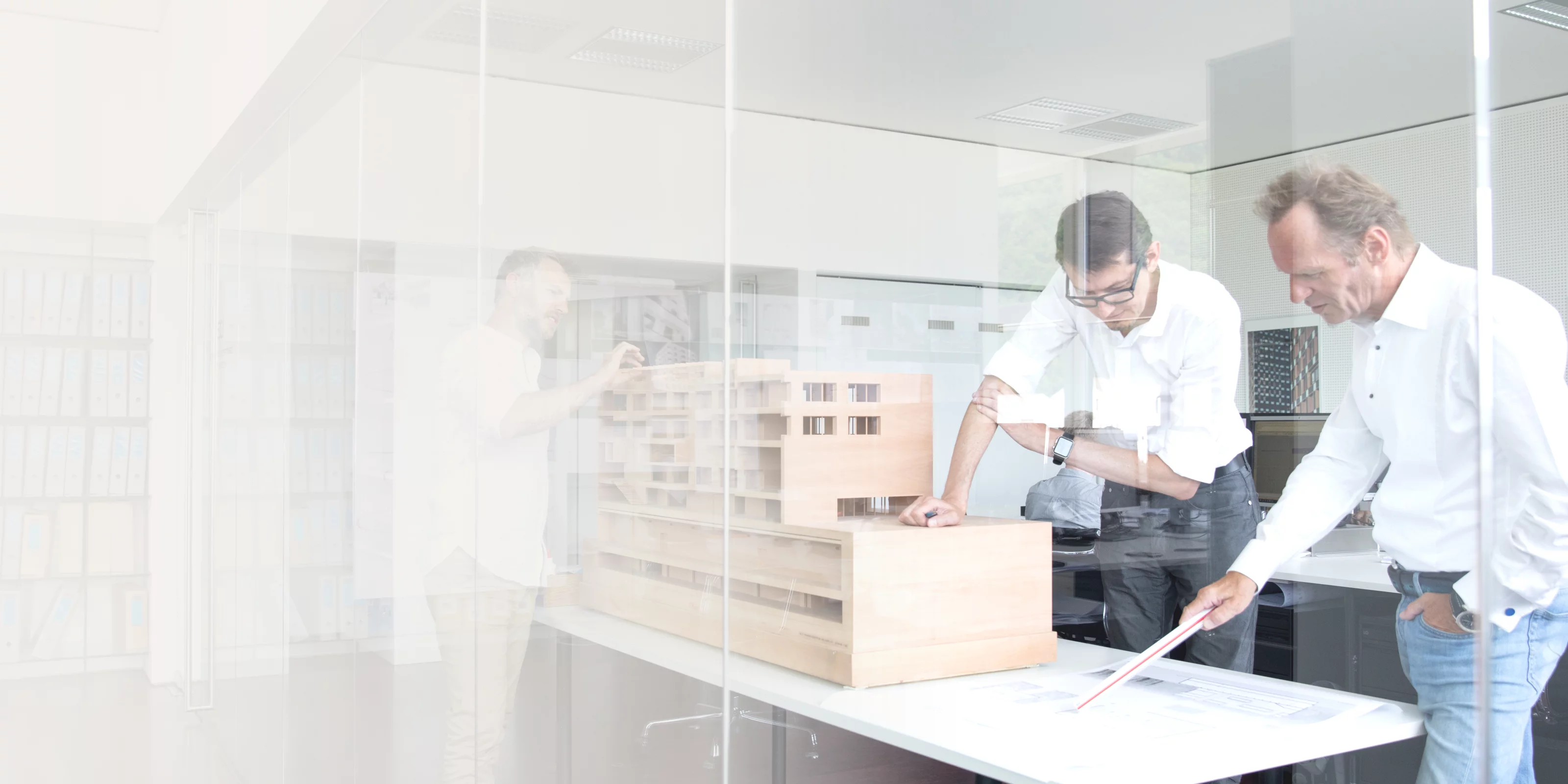 Zwei Personen betrachten ein Architekturmodell auf einem Tisch in einem modernen Büro mit Aktenregalen im Hintergrund.