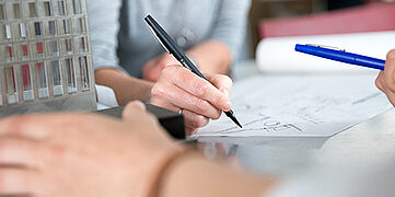 Hände auf einem Tisch, eine mit Stift, der andere mit einem Bleistift. Notizen und ein Modell im Hintergrund.