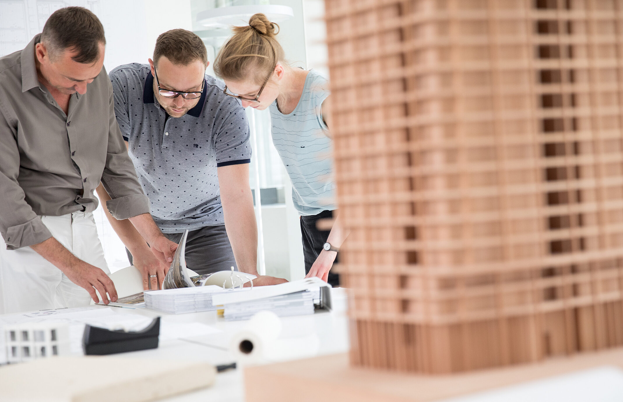 Drei Personen betrachten Pläne und Modelle auf einem Tisch in einem modernen Raum, mit einem Gebäude-Modell im Vordergrund.
