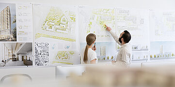 Zwei Personen betrachten Pläne an einer Wand, umgeben von Architekturzeichnungen und Skizzen.