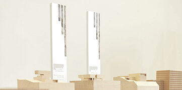 Zwei Auszeichnungen auf Holzsockeln vor einem neutralen Hintergrund, umgeben von architektonischen Modellen.