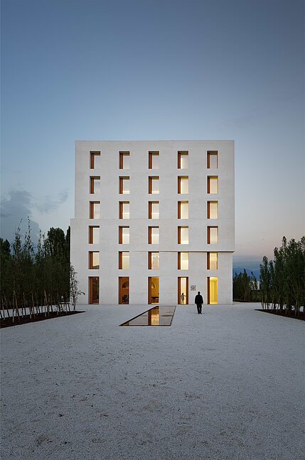 Gegenentwurf zu hochtechnisierten Gebäuden Minimalistisches, weißes Gebäude mit streng rhythmisierten Fensteröffnungen und Vorplatz bei abendlicher Dämmerung